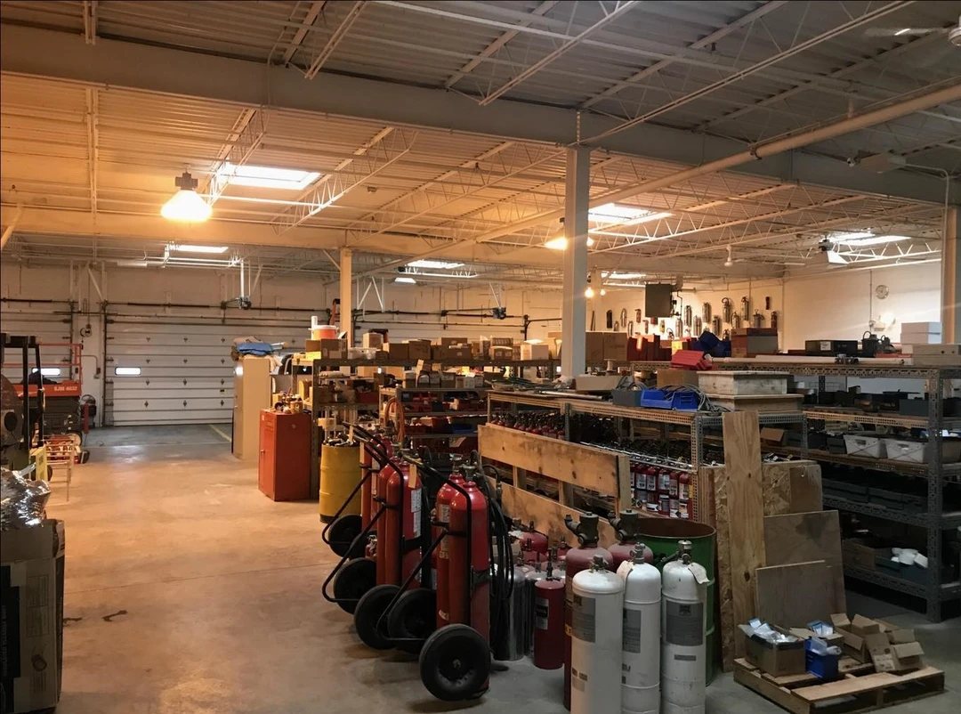 Spacious warehouse interior with shelves stocked with equipment, fire extinguishers, and storage boxes, illuminated by overhead lights